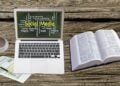 Laptop mit einer Wortwolke rund um Social Media auf einem Holztisch, daneben Kaffeetasse, Wörterbuch und Unterlagen