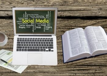 Laptop mit einer Wortwolke rund um Social Media auf einem Holztisch, daneben Kaffeetasse, Wörterbuch und Unterlagen