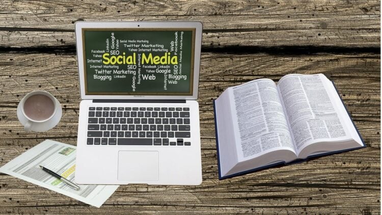 Laptop mit einer Wortwolke rund um Social Media auf einem Holztisch, daneben Kaffeetasse, Wörterbuch und Unterlagen